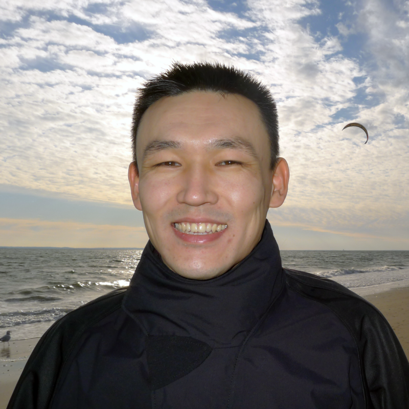 A smiling man in a black jacket stands on a beach with the ocean and a cloudy sky in the background.