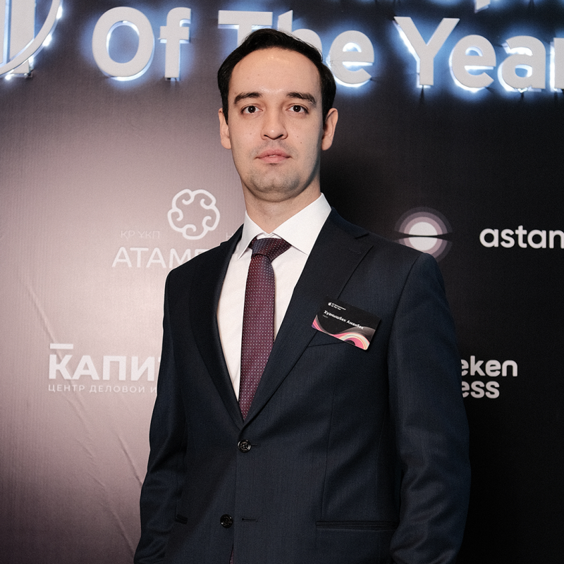 A man in a suit and tie stands in front of a backdrop with logos and the words "Of The Year." He wears a name tag that reads "Куанышбек Аманбек."