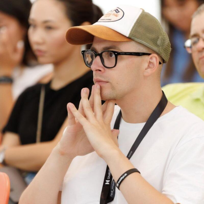 A man wearing glasses and a cap is holding his hands to his face in thought, with a lanyard around his neck and a black bracelet on his wrist.