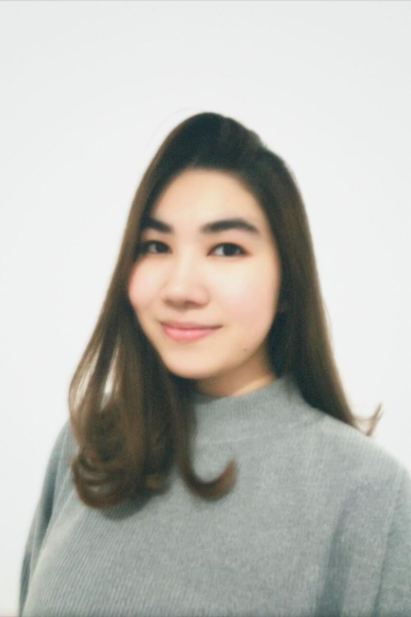 A young woman with long, dark hair smiles in a headshot, wearing a grey turtleneck sweater against a white background.