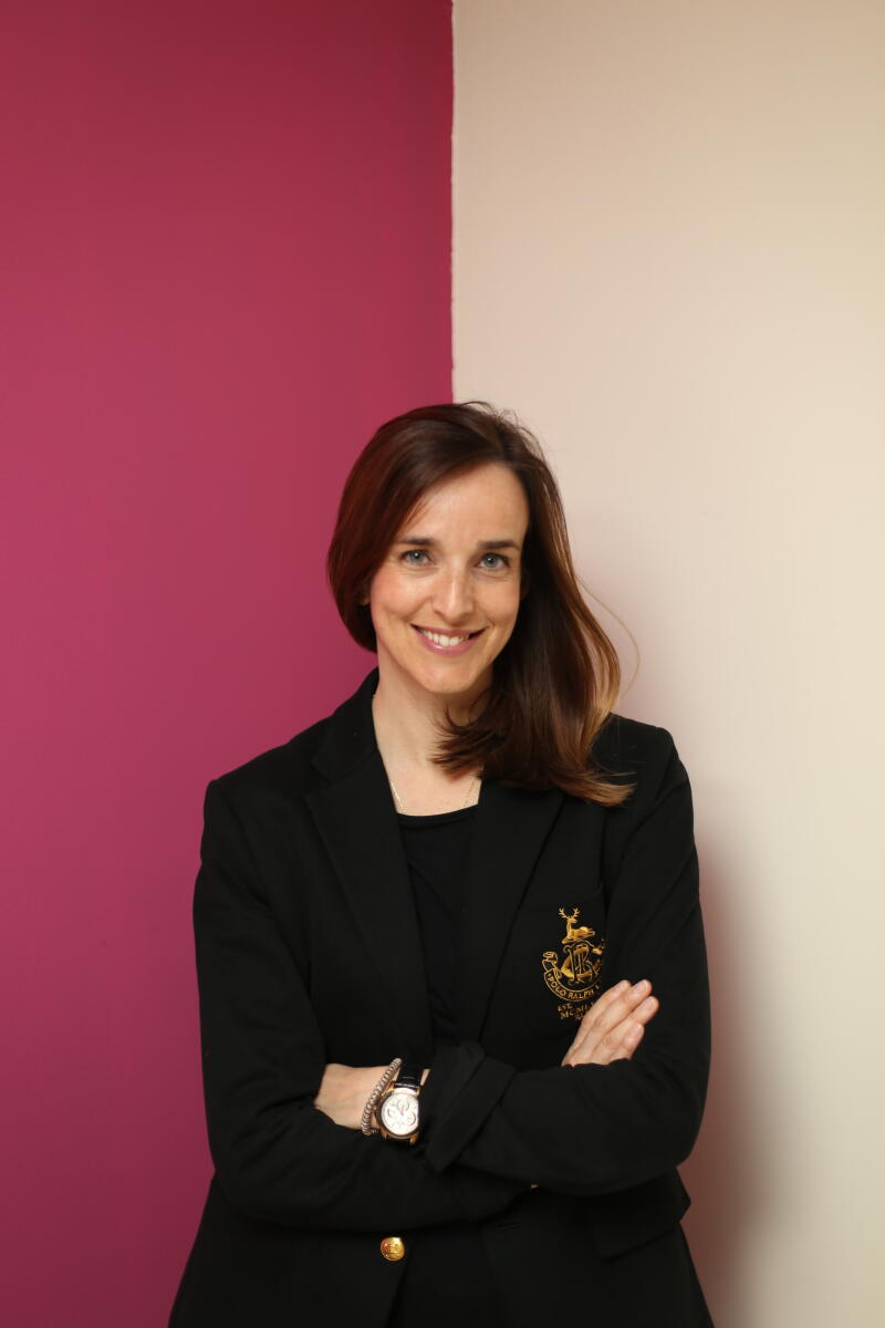 A woman in a black blazer stands with her arms crossed in front of a wall that is half pink and half beige.