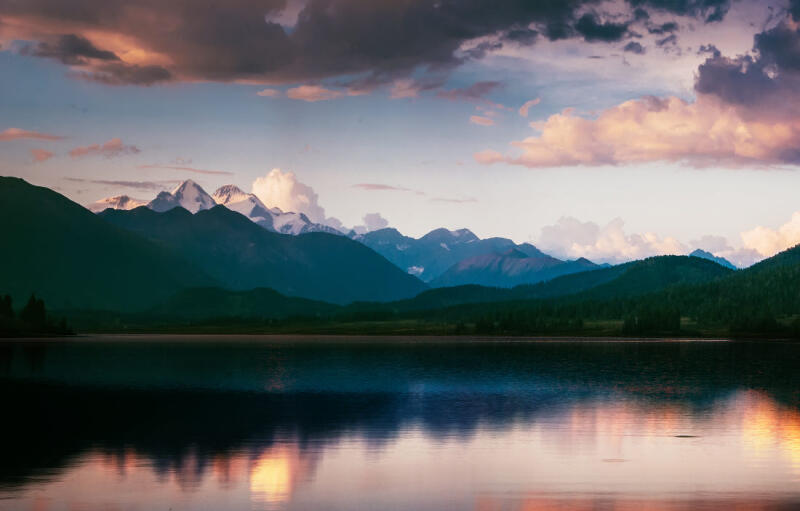 A serene landscape features a calm lake reflecting a mountainous backdrop with snow-capped peaks under a partly cloudy sky.