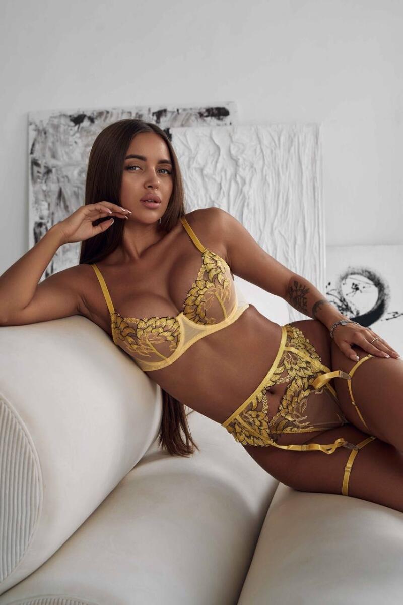 A woman with long brown hair is lying on a white sofa wearing a yellow lingerie set with a floral design.