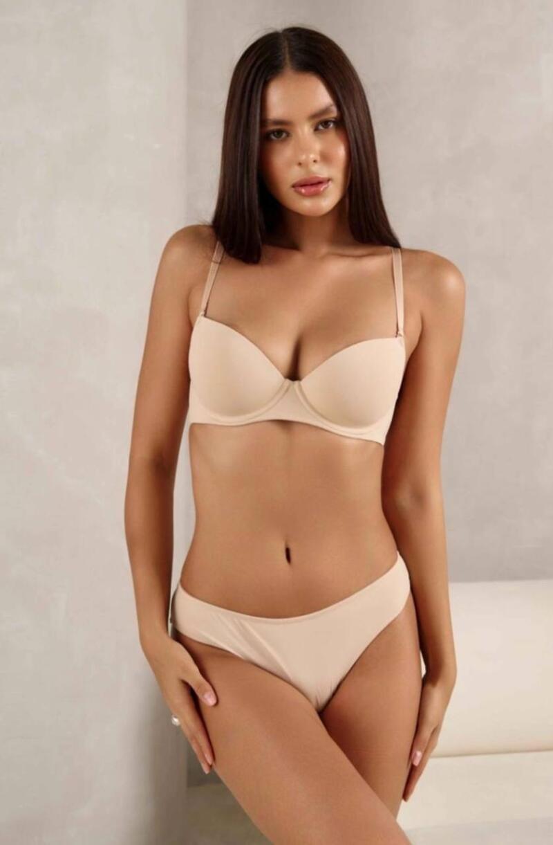 A woman with long brown hair poses in a matching bra and underwear set that is a nude color.