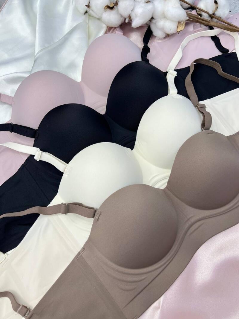 The image shows a pile of strapless bras in various colors: pink, black, white, and beige, with cotton branches in the background.