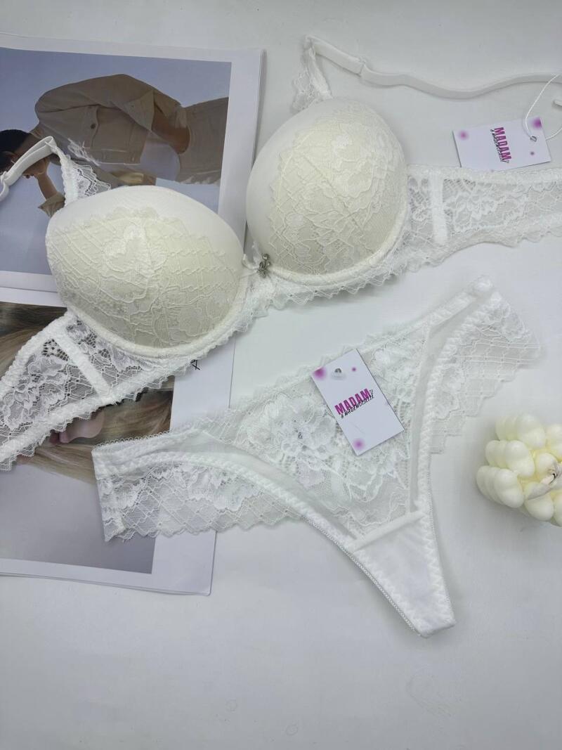 A white lace lingerie set with a bra and thong is laid out on a white surface next to a magazine and small decorative object; the lingerie has tags that say 'MADAM'.