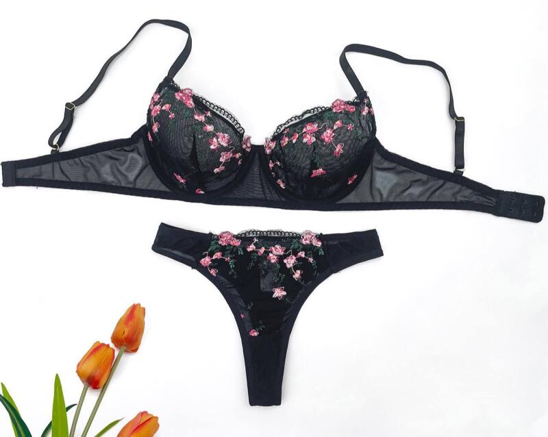 A black bra and panties set with pink floral embroidery, alongside three orange tulips, is arranged on a white surface.