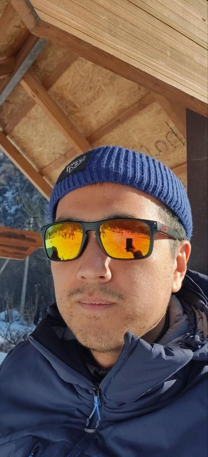 A man in a blue beanie and sunglasses is wearing a blue jacket. The reflection in his sunglasses shows a person holding a phone.