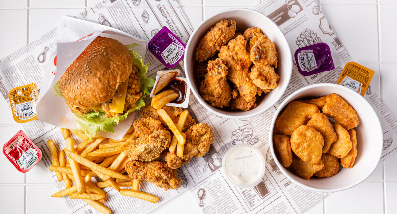 Overhead shot of a burger, fries, chicken wings, and nuggets with various Heinz sauces arranged on newspaper-patterned paper.