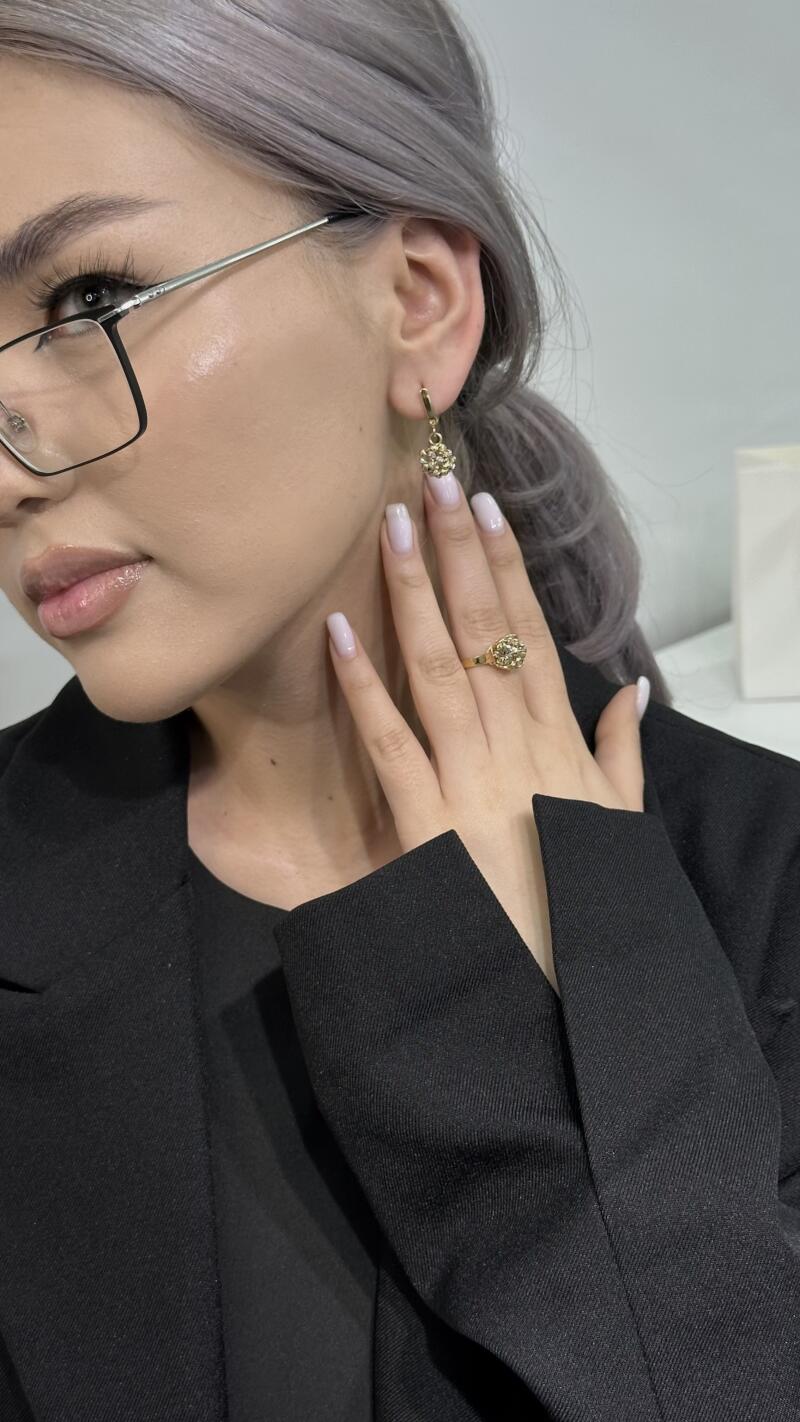 A woman with fair skin, light gray hair, and glasses is wearing a black blazer and gold jewelry, including a ring and earrings.