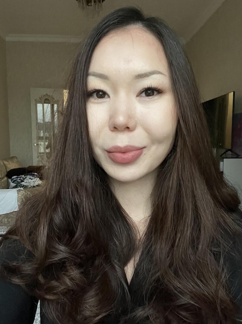 A woman with long, dark hair is looking directly at the camera with a slight smile, wearing minimal makeup.