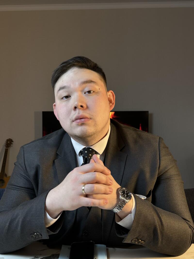 A man in a suit and tie sits at a table with his hands clasped, wearing a watch and a wedding ring.