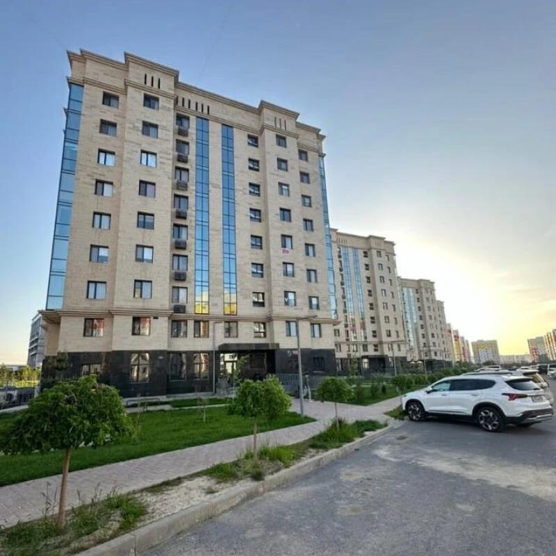 The photo shows a row of multi-story residential buildings with beige facades and blue reflective windows under a clear sky, with a white car parked in the foreground.