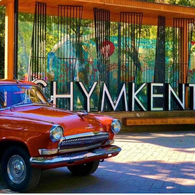 An orange vintage car is parked in front of a structure with the word "SHYMKENT" displayed in large letters.