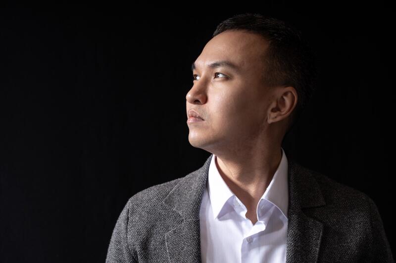 A man in a white shirt and grey jacket looks to the upper right against a black background.