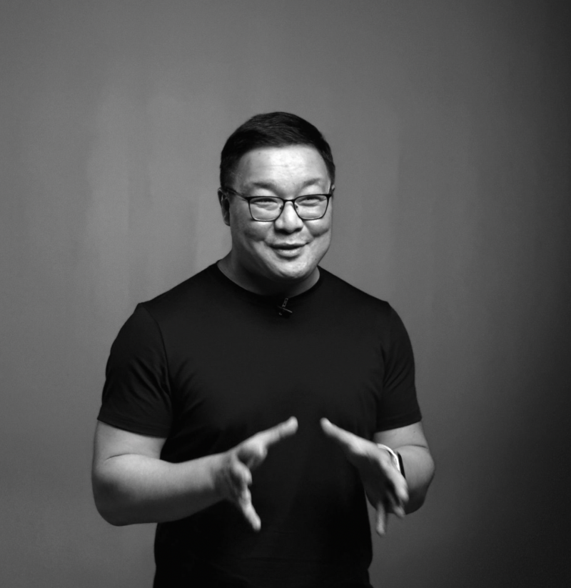 A man with glasses and a black T-shirt stands with his hands gesturing in front of a gray background.