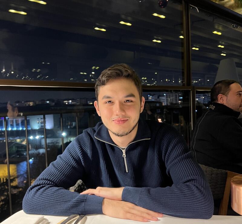 A man with short brown hair and a light beard sits at a table in front of a large window at night, wearing a blue zip-up sweater.