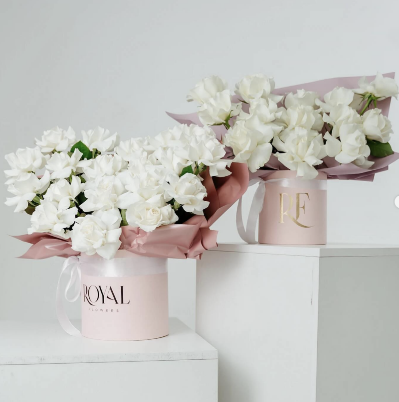 Two arrangements of white roses in pink hat boxes sit on white pedestals, one labeled "ROYAL FLOWERS" and the other "RF."