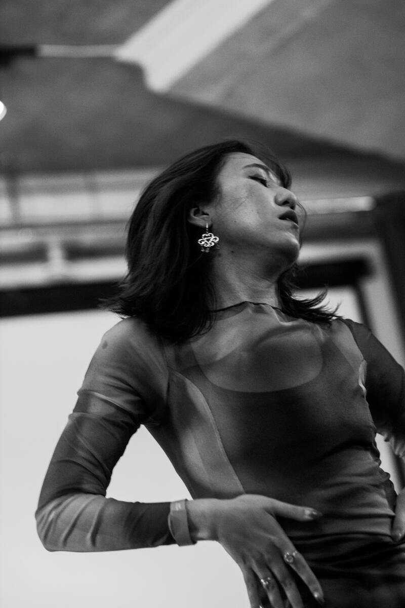 A low-angle, grayscale shot of a woman in a patterned, semi-sheer top with long sleeves, featuring a dark underlayer, and adorned with ornate earrings, with her hand on her hip and her head tilted back.