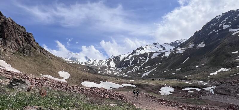A landscape features a mountain valley with snow patches, a dirt path, and two figures walking along it under a cloudy sky.