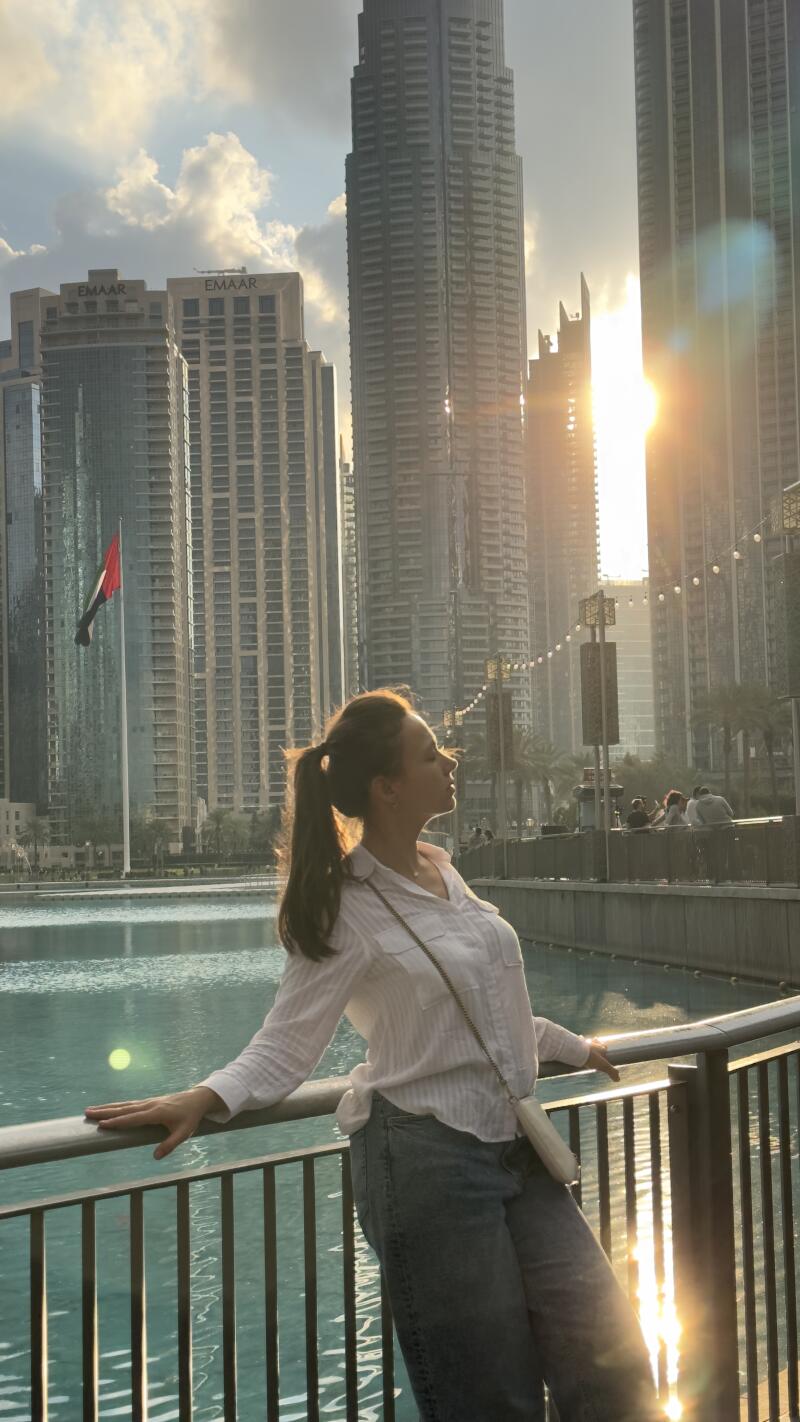 A woman leans against a railing with a pond and skyscrapers in the background on a sunny day, with the word "EMAAR" visible on the buildings.
