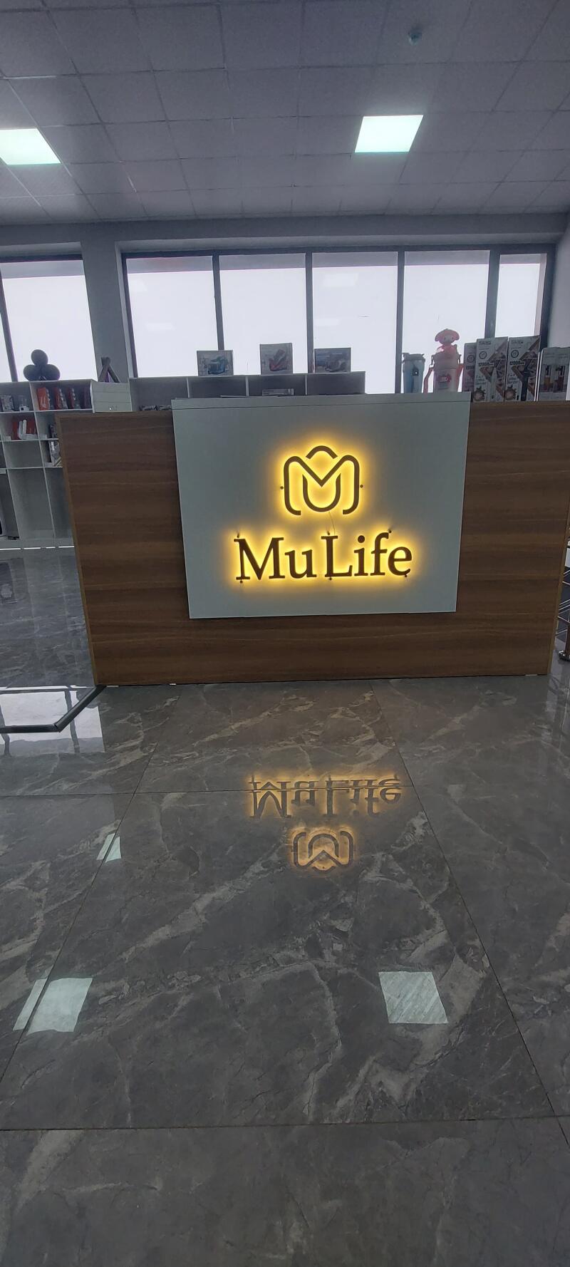 A reception desk with a glowing "MuLife" sign is reflected on a shiny gray marble floor in a brightly lit office space.