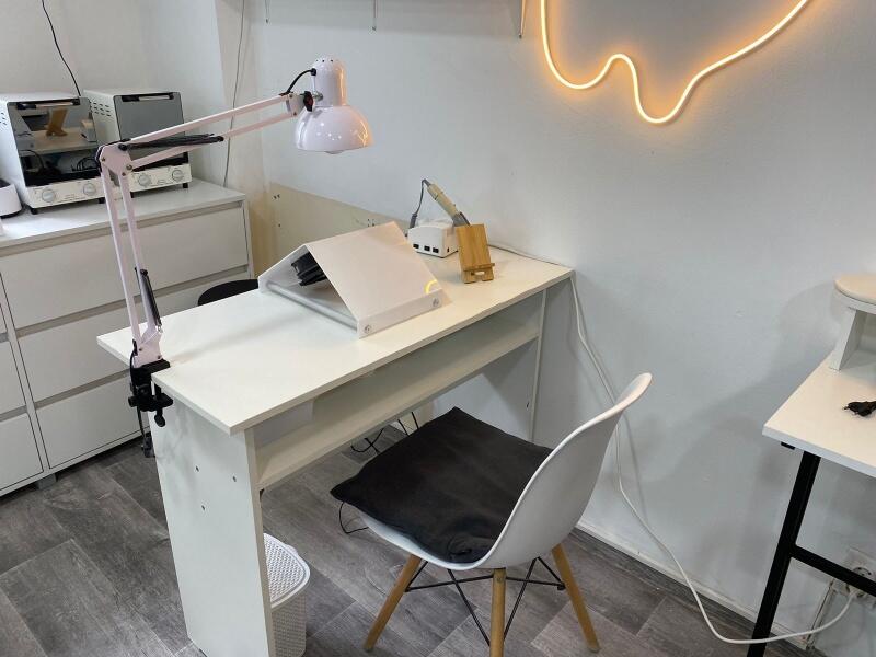 The image shows a white manicure table with a lamp attached to the side and a white chair with a black cushion. On the wall is a neon light in the shape of a drip.