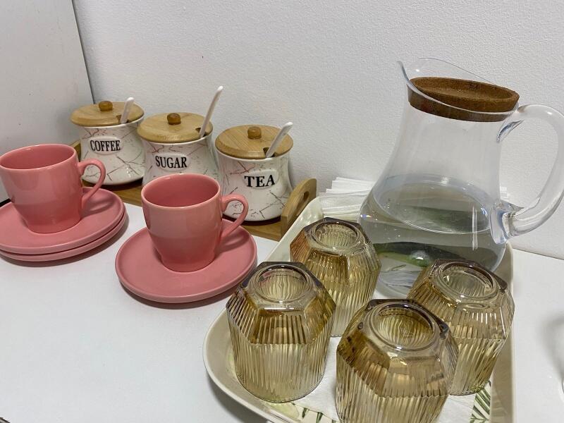 The image shows a set of kitchenware including pink cups and saucers, glass jars labeled "COFFEE," "SUGAR," and "TEA," a clear pitcher filled with water, and several textured brown glasses on a white surface.