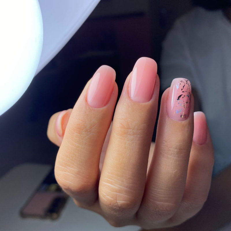 A hand with a set of light pink gel nails, one nail has black and gold decorative elements.