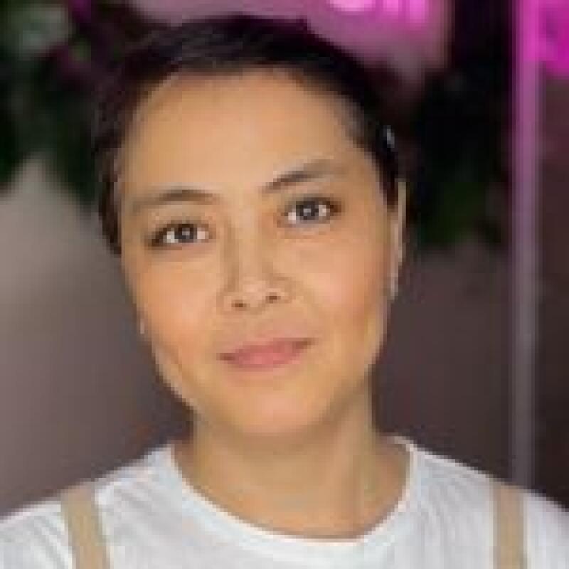A woman with short dark hair, wearing a white shirt, is looking at the camera with a slight smile.