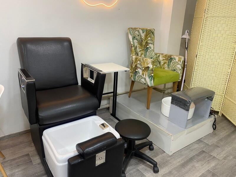 The image shows a pedicure station with a black leather chair with a built-in foot bath, a small black rolling stool, and a chair with a green seat and a leafy pattern on the back.