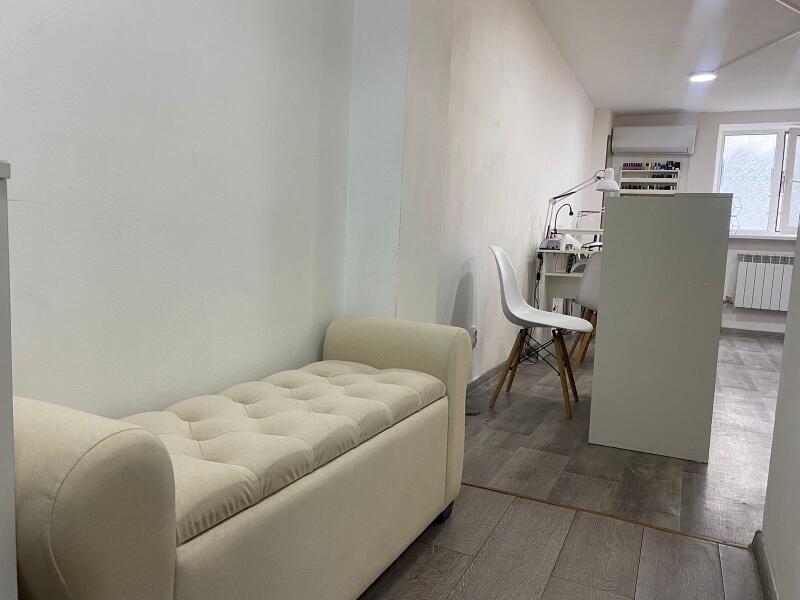 The interior of a room features a cushioned storage bench in the foreground, with white chairs and tables visible further into the space, and a window in the background.