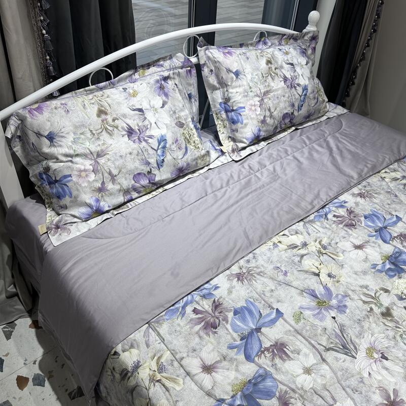 A bed with a white frame is covered with floral-patterned bedding, featuring blue, purple, and white flowers, along with matching pillows and a solid gray comforter.