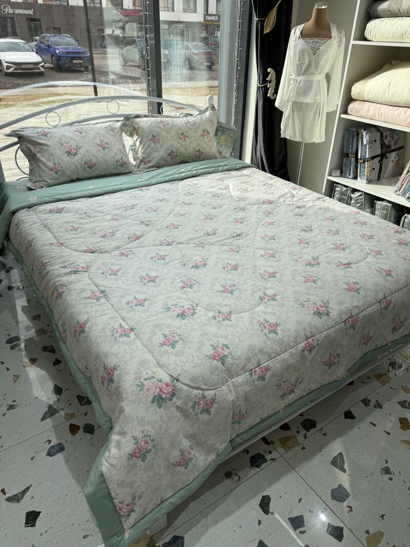 The photo shows a made bed with a floral-patterned comforter and pillows, set against a backdrop of a window and interior furnishings.