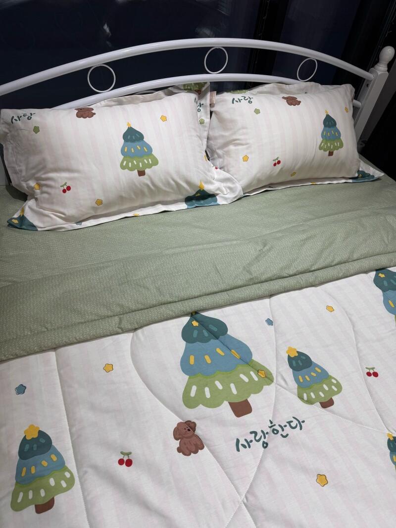 A bed is made with two pillows and a blanket with illustrations of Christmas trees, cherries, a dog, and the Korean text "사랑한다."