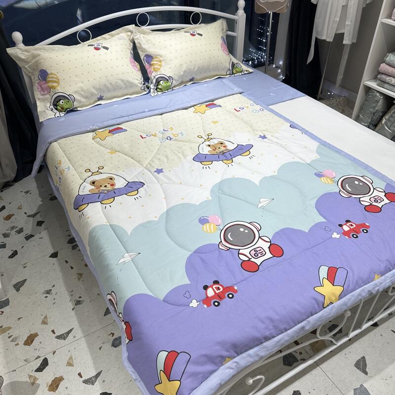 A bed is covered with a colorful, cartoon-themed comforter and pillows, featuring images of astronauts, bears in UFOs, cars, and stars.