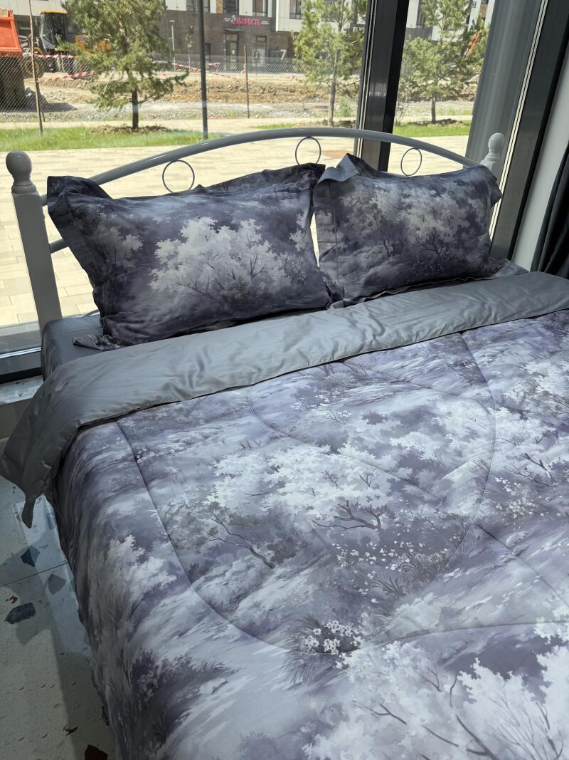 A bed with grey patterned bedding and pillows, featuring a design resembling a forest landscape, sits near a window.