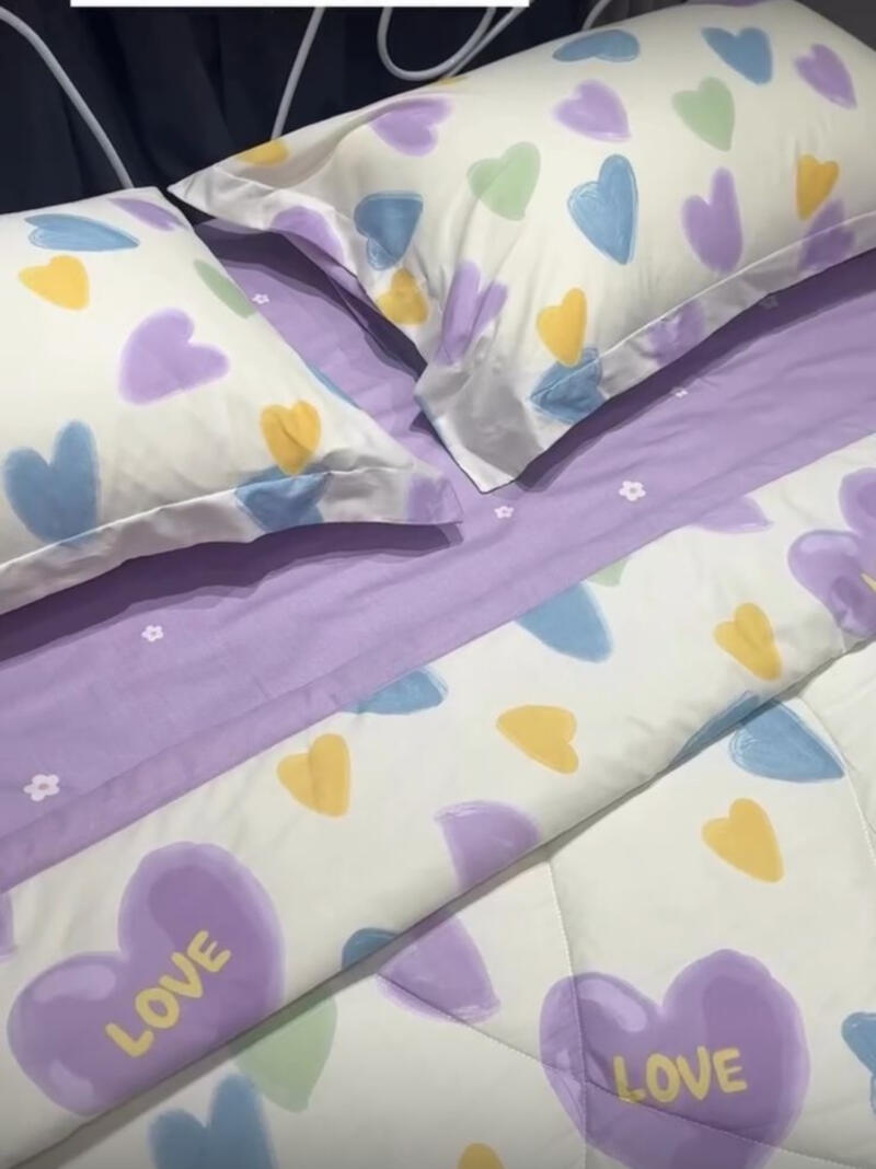 A bed is covered with a white sheet and pillows, all patterned with colorful hearts; some of the hearts contain the word 'LOVE'. The sheet has a lavender band across the middle.