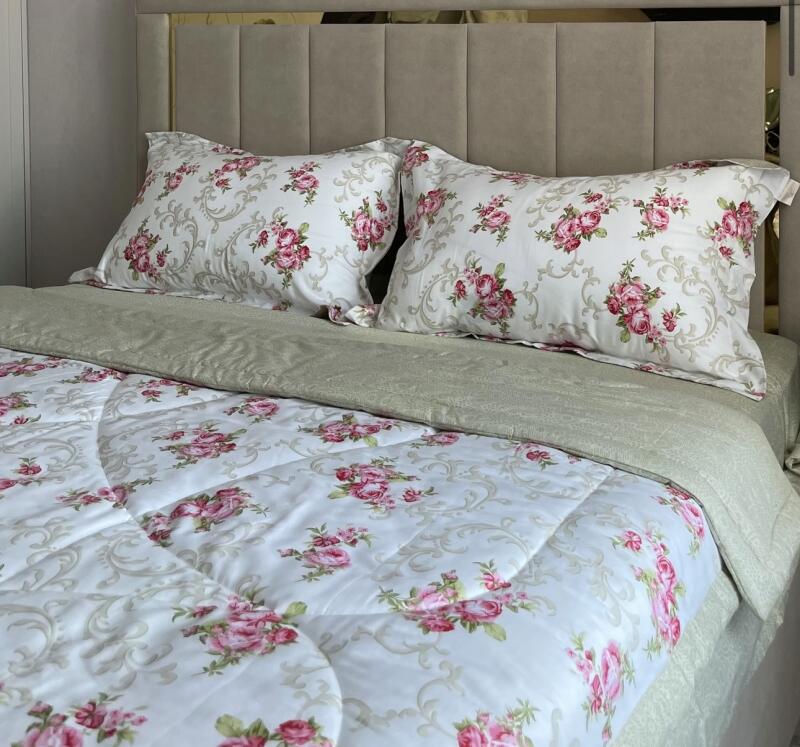 A bed with a beige headboard is covered with a light green sheet and white bedding patterned with pink roses and beige swirls, with two matching pillows.