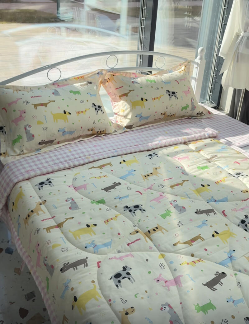 A bed with a white metal frame is covered with bedding featuring a pattern of colorful dogs, topped with two pillows with a similar dog motif.