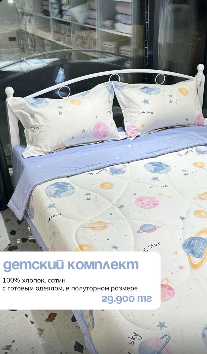 A bed with white metal frame and a set of bedding with a space theme including planets and stars, with text overlay reading "детский комплект 100% хлопок, сатин с готовым одеялом, в полуторном размере 29.900 тг"