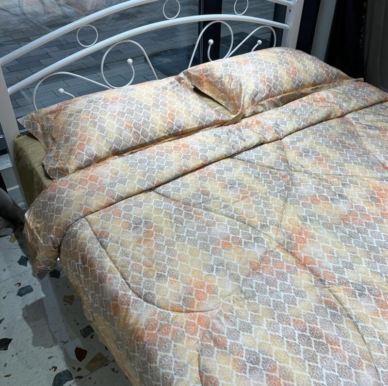 A bed with a white metal frame is covered with a patterned beige, orange, and gray comforter and pillows.