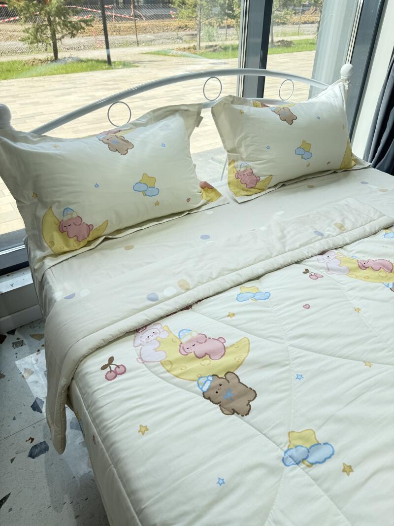 A bed with a white metal frame is covered with a light yellow comforter and pillows, all patterned with cartoon bears on moons, stars, and clouds.
