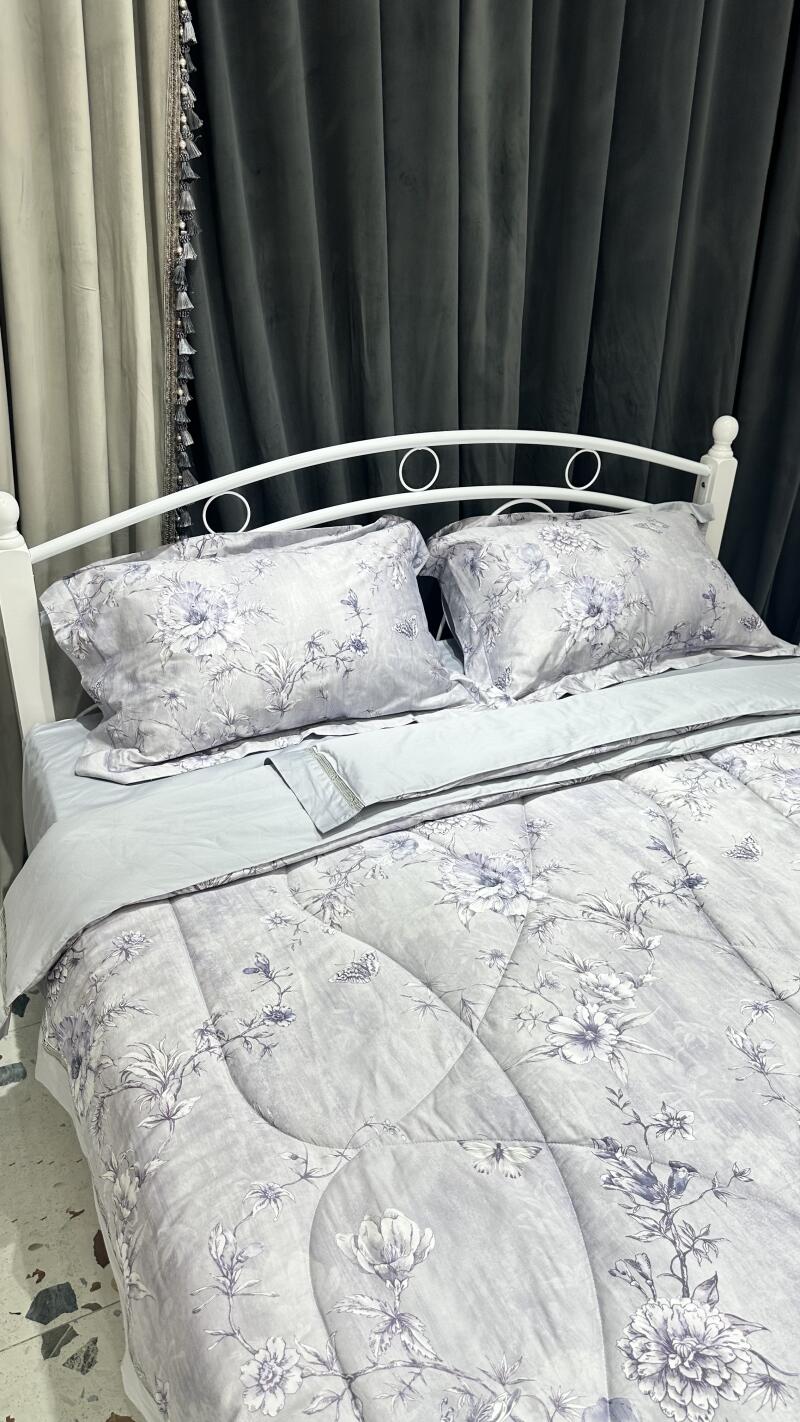 A bed with white metal frame is covered in light blue floral bedding, with matching pillows, set against a backdrop of gray curtains.