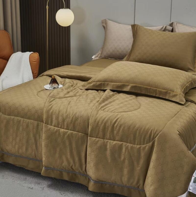 A tan comforter with a subtle repeating pattern is neatly laid out on a bed with matching pillows, and a small bottle sits on a saucer on top of the comforter.