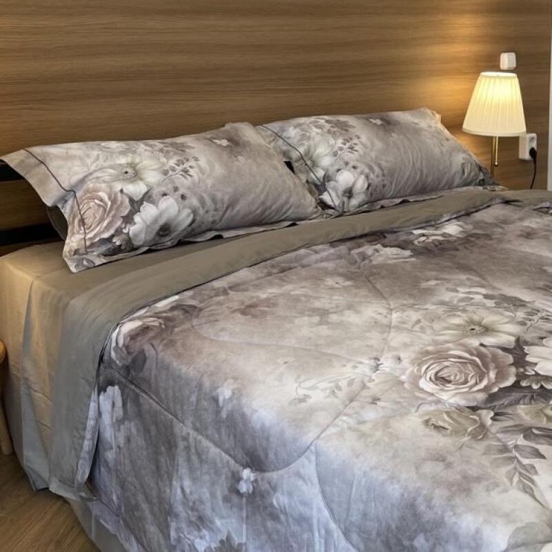 A neatly made bed features pillows and a comforter with a grey floral pattern, set against a wooden headboard with a lamp attached to the wall.