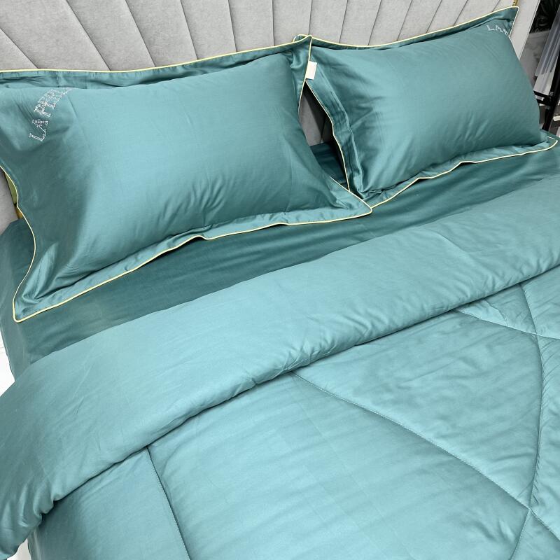 A bed is neatly made with two matching pillows and a comforter, all in a teal color with a subtle sheen. The pillows feature a delicate embroidered "LA VIE" design, and a thin yellow piping borders the edges of the bedding.