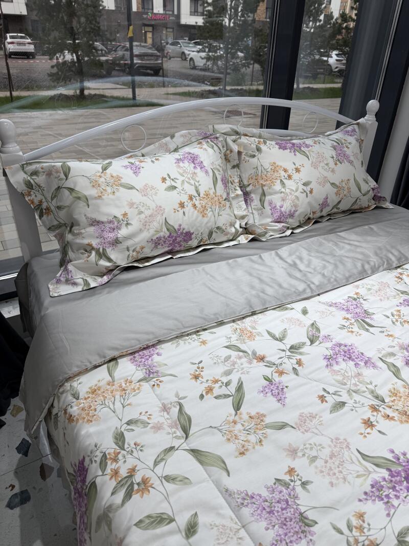 A bed with a white frame is covered with a grey sheet and a white comforter and pillows with a floral pattern in purple and orange.