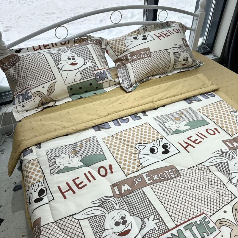 A bed is covered with a cartoon-themed comforter and pillows featuring images of cats and rabbits, along with text such as "HELLO!" and "I'M SO EXCITED."