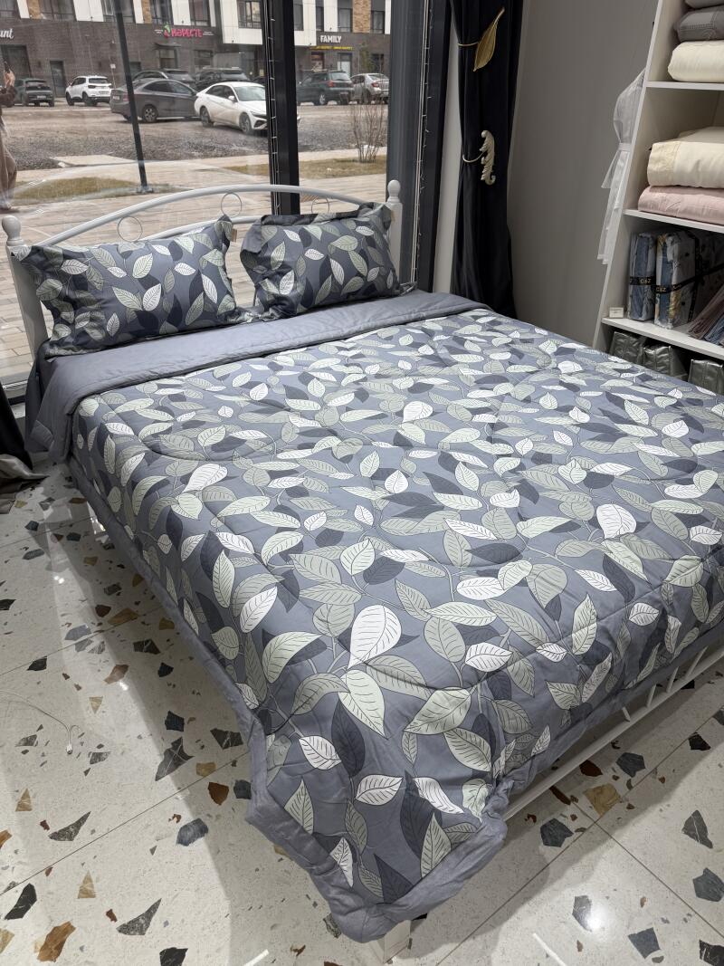 A bed with gray leaf-patterned bedding and two matching pillows is set up in front of a window.